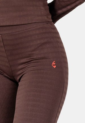 Close-up van bruine, getextureerde leggings met een klein rood geborduurd symbool met een hart op de bovenste dij.