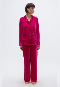 Γυναίκα όρθια, ξυπόλυτη, φορώντας ένα κουμπωτό ματζέντα βελούδινο σετ πιτζάμας με μακριά μανίκια και παντελόνια φαρδιά, κοιτώντας προς τα αριστερά της.