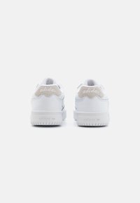 Ett par vita Adidas-sneakers sedd från baksidan med en beige mocka hälflik och Adidas-loggor på hälen och sulan.