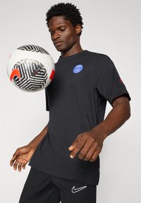 Sort kortærmet t-shirt med blåt logo, båret af en person, der holder en sort-hvid mønstret fodbold med røde accenter.
