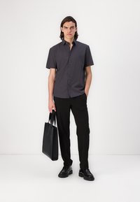 Kortermet skjorte med mørkt mønster, kombinert med svarte bukser og sko. Modellen holder en svart tote-veske og står foran en ensfarget bakgrunn.