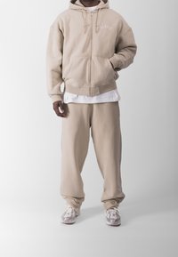 Sudadera beige con cremallera, bolsillos frontales y cordón, combinada con pantalones jogger a juego. Zapatillas ligeras completan el conjunto.