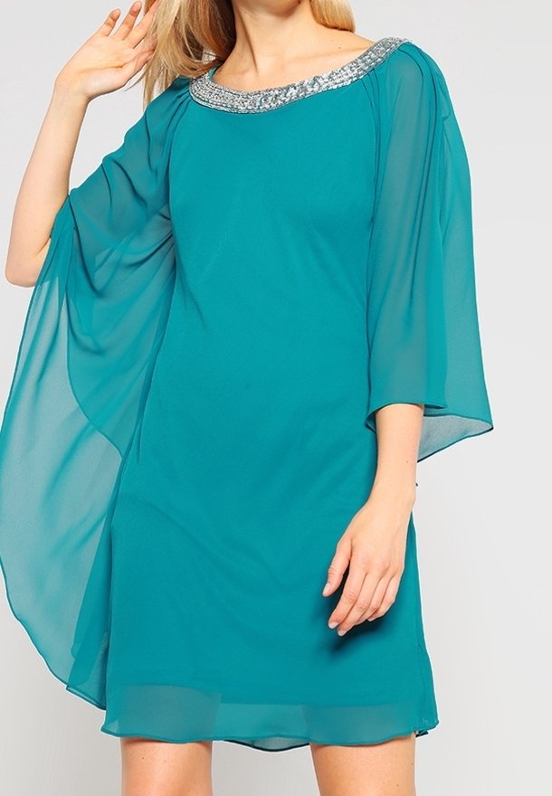 Femme portant une robe turquoise longueur genoux avec des manches transparentes et fluides et un décolleté orné de perles argentées, posant avec une main levée près de la tête.