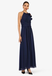 Marineblaues, langes Kleid aus fließendem Stoff mit einem Halterneck, plissierter Brustpartie und floralem Akzent an einer Schulter. Schwarze High Heels.
