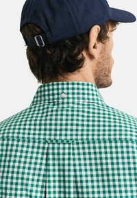 Groen en wit geblokt shirt met een button-down kraag en een rugplooidetail, gedragen met een donkerblauwe pet.