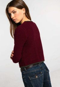 Suéter burdeos texturizado con un diseño corto y cuello redondo, usado sobre una blusa oscura. Combinado con jeans de mezclilla oscuros con bolsillos.