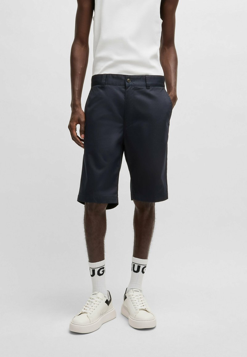 Mörkblå shorts med platt front, sidfickor och bältesöglor. Kombineras med en vit ärmlös topp och vita sneakers med svarta detaljer.