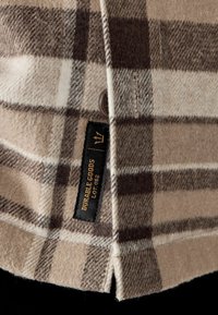 Beżowa i brązowa tkanina w kratę o miękkiej teksturze. Posiada czarną metkę z białym i złotym napisem oraz brązowy guzik na krawędzi.