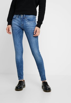 Person, die ausgewaschene blaue Skinny-Jeans, ein schwarzes langärmliges Oberteil und schwarze Schnürschuhe trägt und vor einem schlichten hellen Hintergrund steht.
