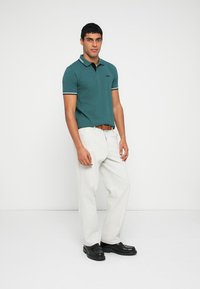 Žalsvai mėlynas polo marškinėlis su dvikoliu apykaklės raštu, derinamas su baltomis kelnėmis ir juodais batais. Pasižymi liekninančiu dizainu ir kontrastinėmis siūlėmis.