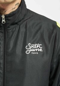 Svart zip-up jakke med ribbet tekstur, lysende gule detaljer på skuldrene, og "Sixth June Paris"-logo i hvitt.
