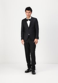 Čierny smoking so saténovým špicatým klopom, biela košeľa a čierna motýliková kravata. Doplnené lesklými čiernymi dress shoes, stojí na jednoduchom pozadí.