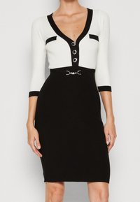 Svart och vit klänning med V-ringning, trekvartslånga ärmar, knappar framtill och en metallisk detalj vid midjan.