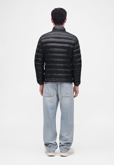 Homme portant une veste matelassée noire, un jean bleu clair ample et des chaussures blanches, debout de dos sur un fond uni.