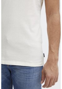 Creme bomulds T-shirt med korte ærmer, med en ensfarvet tekstur og et lille mærke på siden. Kombineret med blå denim jeans.