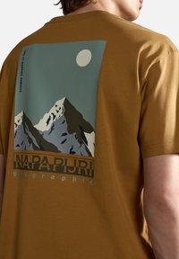 Camiseta de algodón marrón con un gráfico de montañas y una luna en la parte posterior. Texto: "NAPAPIJRI geographic." Mangas cortas y corte relajado.
