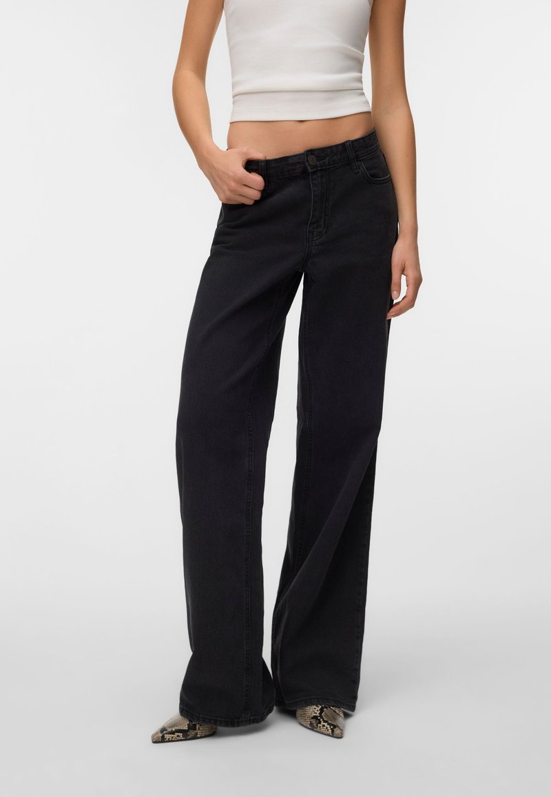Femme vêtue d’un jean noir à jambes larges, d’un débardeur blanc court et de chaussures pointues à motif peau de serpent, debout sur un fond uni.