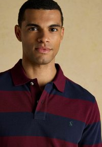 Polo krekls ar bordo un tumši zilām horizontālām strīpām, izgatavots no kokvilnas materiāla, ar diviem pogām pie apkakles, rotāts ar mazu zaļu logotipu uz krūtīm.