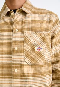 Brun og creme ternet flannel skjorte med knaplukning, en lomme på venstre side og et logo-patch med mærket "Dickies."
