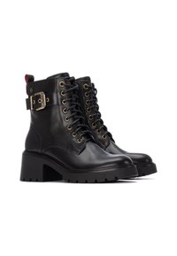 Botas de cuero negro hasta el tobillo con un tacón grueso, parte delantera con cordones, ojales dorados y detalle de hebilla en el lado. Suela de goma texturada.