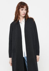 Long cardigan gris foncé en maille douce. Il a un col en V, une ouverture devant, et deux poches avant avec des détails côtelés.