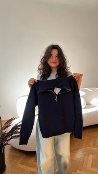 Tengerészkék kötött pulóver, negyedzipzárral, bordázott textúrával és felgöngyölt gallérral, egy világoskék nadrágot viselő személy kezében.