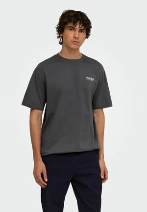 Giovane uomo con capelli ricci scuri che indossa una maglietta grigio scuro con il testo "modul" sul petto e pantaloni scuri contro uno sfondo semplice.
