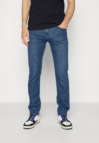 Modré džíny z denimu s rovnými nohavicemi, mají pět kapes a viditelné švy. Nosené s černým tričkem a barevnými teniskami.