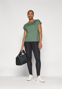 T-shirt verde oliva a maniche corte abbinato a leggings neri e sneakers bianche. Borsa a tracolla nera con manici. Look di abbigliamento sportivo semplice e funzionale.