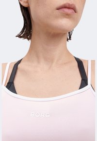 Κοντινό πλάνο ενός ατόμου που φοράει ανοιχτό ροζ αμάνικη μπλούζα με λευκό περίγραμμα και ορατές μαύρες τιράντες αθλητικού σουτιέν από κάτω.