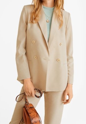 Femme portant un blazer croisé beige et un pantalon, tenant un sac à main marron à motif peau de serpent, avec des colliers en or superposés.