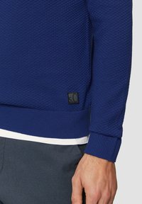 Granatowy sweter o fakturowanej strukturze z prążkowanymi mankietami i dołem. Posiada małą czarną metkę z napisem „s.O” na dolnej lewej stronie.