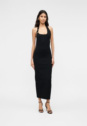 Femme portant une robe midi noire sans manches à col licou avec une boucle argentée et des sandales noires à talons hauts, debout devant un fond uni.