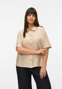 Kortærmet, kravebluse i lys beige stof, med knaplukning foran og en afslappet pasform. Sammen med mørke denimbukser.