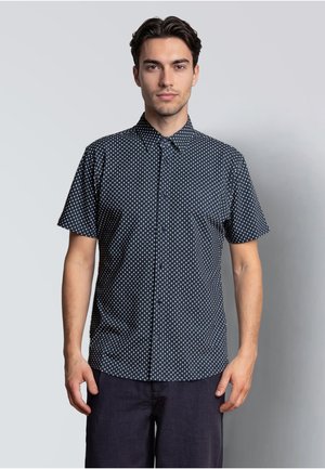 Mann steht vor einfarbigem Hintergrund, trägt ein navyblaues Kurzarmhemd mit weißen geometrischen Mustern und dunkle Hosen.