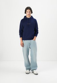 Navy hoodie with drawstrings and front pocket, paired with light blue baggy jeans and white sneakers with black accents.