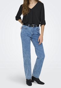 Blouse noire à col en V avec manches retroussées, associée à un jean en denim bleu taille haute et des bottines noires, agrémentée d'une ceinture noire brillante.