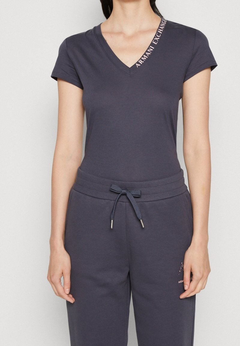 Femme portant un t-shirt gris foncé à manches courtes en V avec le texte "Armani Exchange" sur le col et un pantalon assorti à taille élastique avec cordon de serrage.