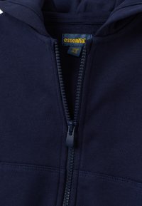 Sudadera con cremallera azul marino, hecha de un tejido suave, con una cremallera de contraste, capucha y una etiqueta interior que muestra "esencial". Textura suave.