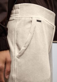 Pantalons en velours côtelé beige clair avec une texture côtelée, dotés d'une poche latérale et d'une petite étiquette sombre près de la taille. La ceinture est épaisse et texturée.