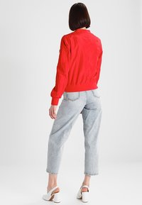 Veste bomber rouge avec poignets et ourlet élastiques, associée à un jean taille haute bleu clair et des sandales blanches à talons blocs.