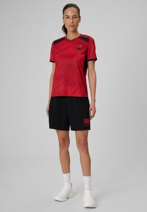 Kobieta stoi, ubrana w czerwoną sportową koszulkę z czarnymi detalami, czarne szorty, białe skarpetki i białe buty sportowe na jednolitym szarym tle.