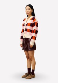 Vrouw die staat in een roze, oranje en bordeauxrode geruite cardigan, bordeauxrode leren rok, bordeauxrode sokken en bruine fluffy schoenen.