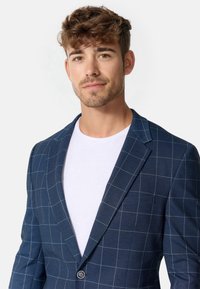 Marineblå ternet blazer lavet af tekstureret stof, med en enkelt knaplukning og klapkrave, båret over en hvid t-shirt.