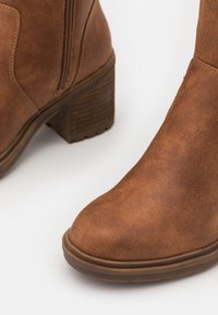 Brązowy botek za kostkę z sztucznej skóry, z okrągłym noskiem, teksturowaną powierzchnią, bocznym zamkiem i grubym obcasem. Wzmocniona guma podeszwy.