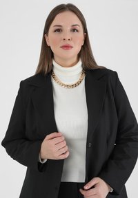 Sort blazer over en hvid ribbet polokrave, suppleret med et chunky guld kædedesign, som giver et struktureret og poleret udseende.