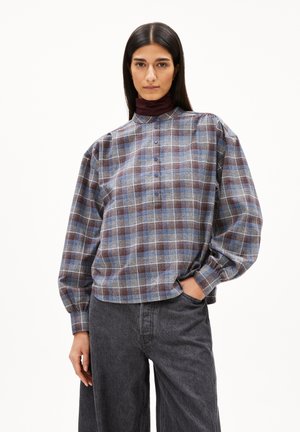 Ruitjesoverhemd in blauw, bruin en wit. Heeft een hoge kraag, knoopsluiting en losse mouwen. Gecombineerd met donkergrijze broek.