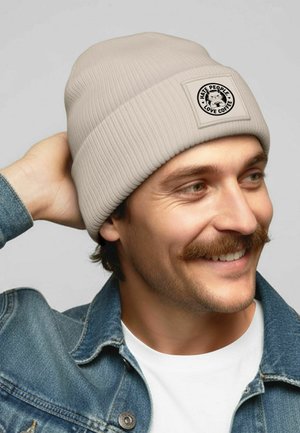Smiling man with mustache wearing beige beanie with a cat patch saying "Hate People, Love Coffee," denim jacket, and white shirt.
