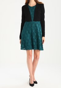 Teal Spitzenkleid mit eng anliegendem Oberteil und schwingendem Rock, kombiniert mit einer schwarzen cropped Strickjacke. Verfügt über kurze Ärmel und einen gewellten Saum.