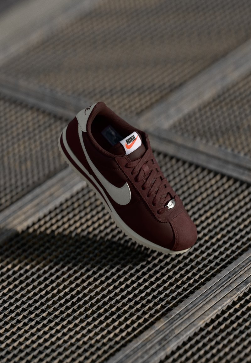 Sneaker Nike con una parte superiore color bordeaux, logo swoosh crema, chiusura con lacci e suola bianca, posizionata su una superficie testurizzata.